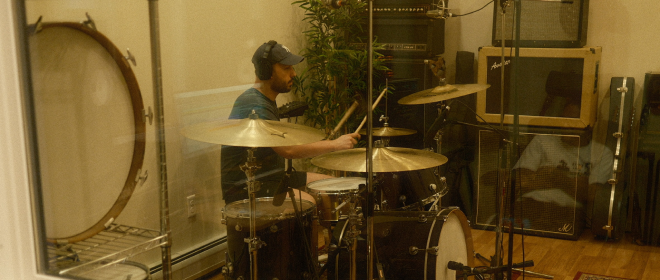 Drummer recording in a soundproof studio room, surrounded by microphones, drums, and vintage amplifiers.